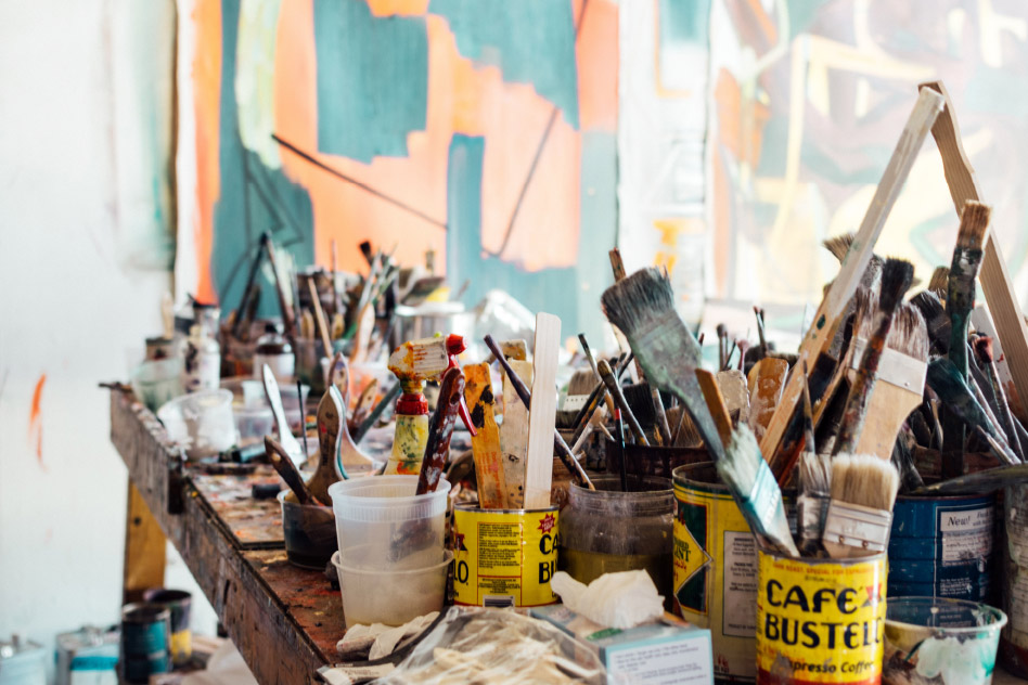A messy table in an artist studio covered with cans of paint, brushes, and assorted art supplies